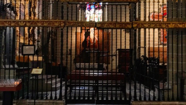 Interior de una iglesia en penumbra visto a través de una reja de hierro ornamentada, con una vidriera iluminada al fondo